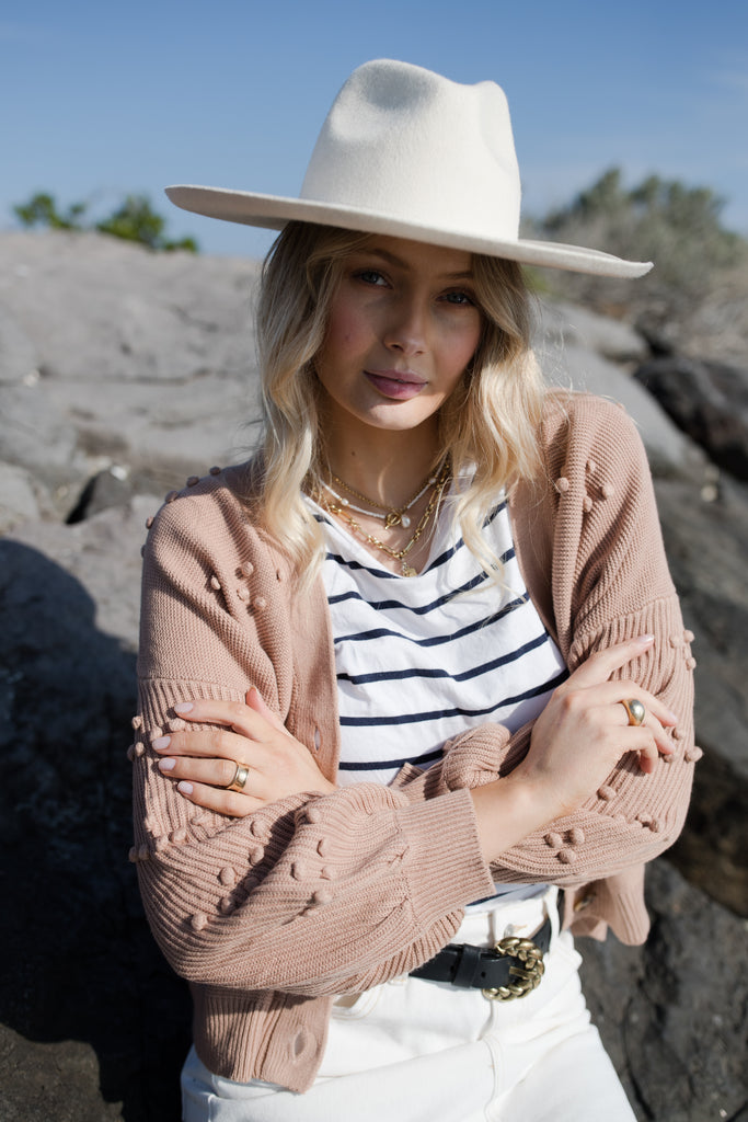 Rhiannon wears the Bowie Bobble Knit cardigan in Sand, she is wearing a White felt hat, striped tank and White denim midi skirt with a belt.